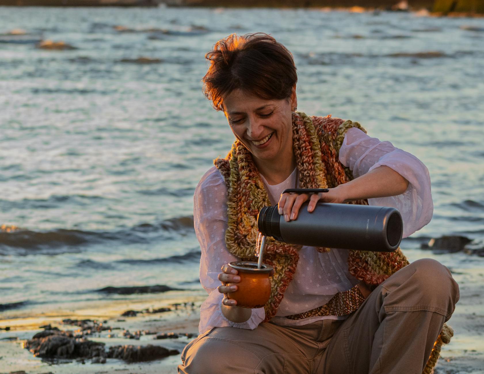 Kobieta siedzi nad morzem o zachodzie słońca i pije napar z mate