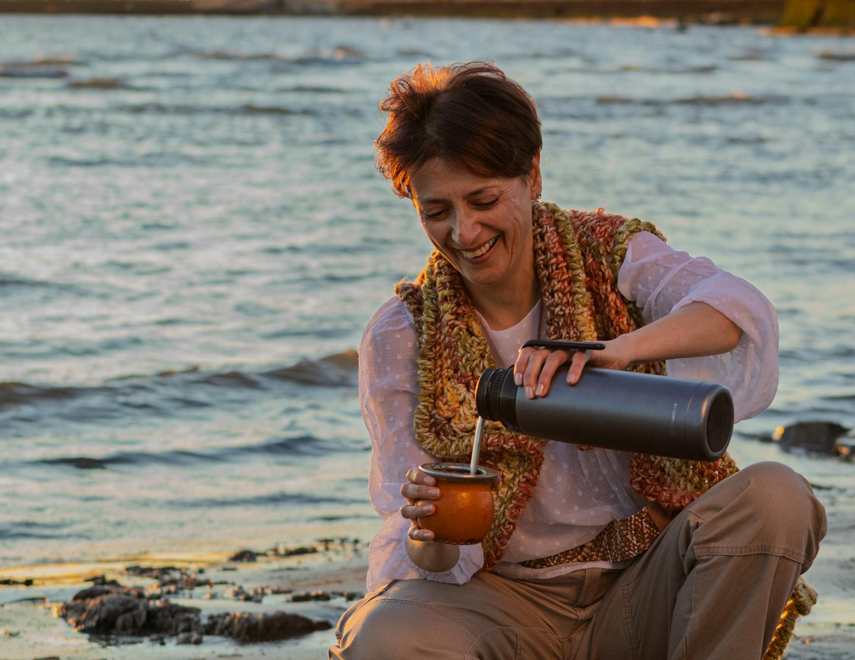 Uśmiechnięta kobieta pije napar z termosu na plaży o zachodzie słońca